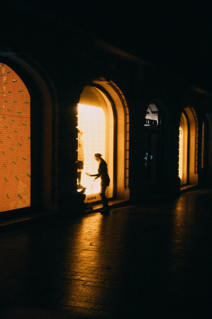 Silhouette of a person by lit shop windows in an arched building at night.